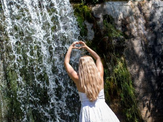 Frau vor Wasserfall haltet die Hände zu einem Herz geformt 