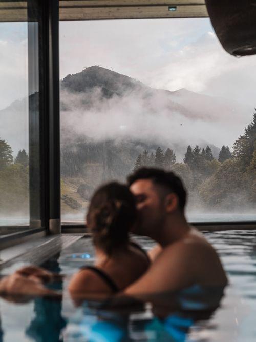 Stimmungsvolles Bild einens Paares, welches im Pool schwimmt und dahinter ein ausblick in die Grüne Berglandschaft 