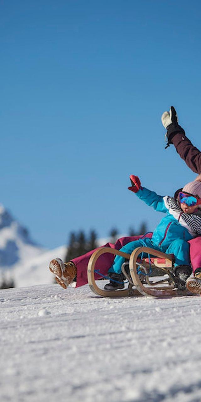 Mutter und Kind rodeln einen verschneiten Hang hinunter vor Alpenkulisse