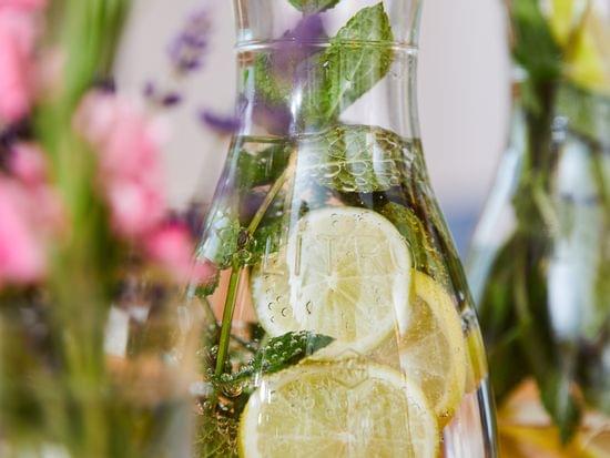 Glass carafe with lemon slices, mint leaves, and sparkling water among flowers