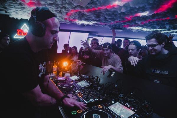 DJ vor seinem Pult, davor Gäste die Tanzen - Club Stimmung, beleuchtete Decke 