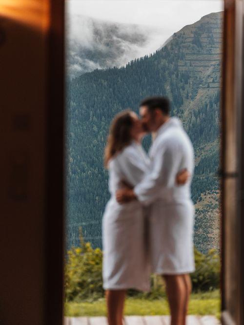 2 Personen im Bademantel, die sie umarmen und sich küssen, im Hintergrund die Berge 