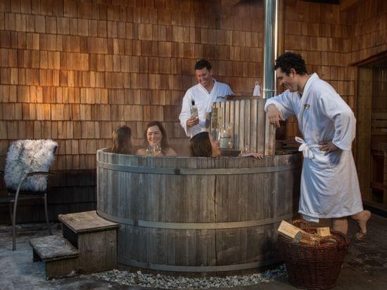 Freunde entspannen im hölzernen Außen-Whirlpool mit Wein und Bademänteln