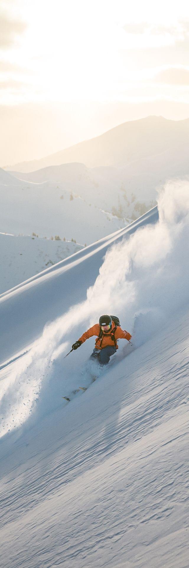 Skifahrer in orangefarbener Jacke fährt durch tiefen Pulverschnee am sonnigen Berghang