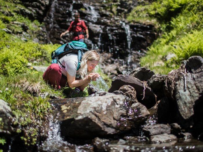 Frau trinkt frisches Bergquellwasser beim Wandern in einer grünen Alpenlandschaft