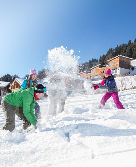 Gruppe von Menschen beim Schneeballwerfen vor Alpenhütten an einem sonnigen Wintertag