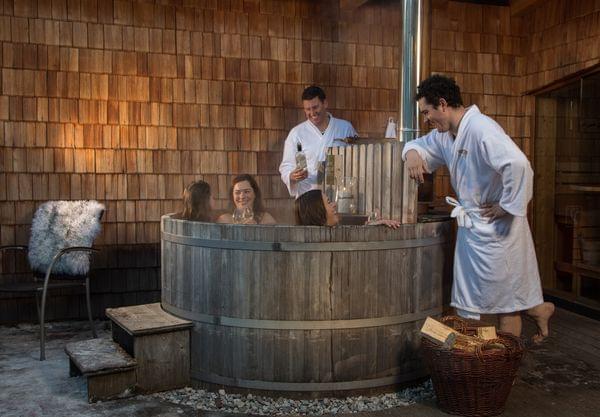 Freunde entspannen im hölzernen Außen-Whirlpool mit Wein und Bademänteln