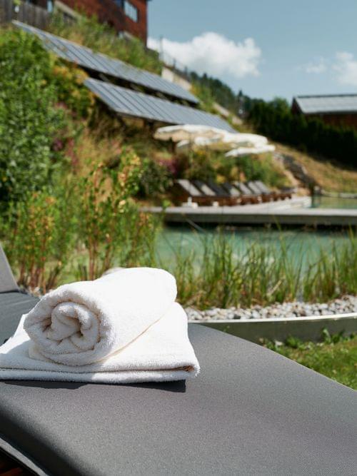 Wellnessliege mit weißem Handtuch neben Naturpool in sonnigem Garten