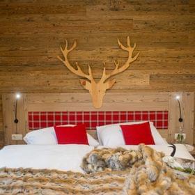 Gemütliches Hotelbett mit Felldecke, roten Kissen und Hirschkopf aus Holz an rustikaler Holzwand