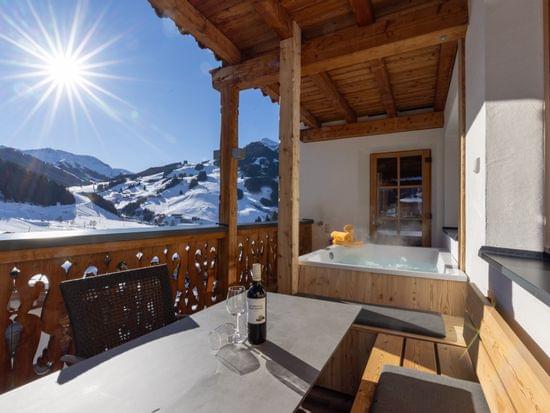 A terrace with a dining table and chairs and a hot tub, overlooking a snowy winter landscape in bright sunshine.