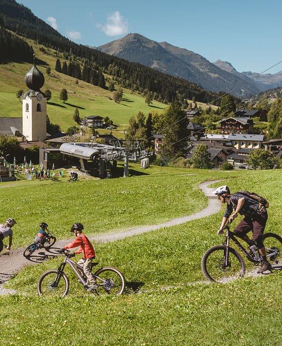 Familie beim Mountainbiken auf einem Trail im Alpendorf mit Seilbahn und Bergen im Hintergrund