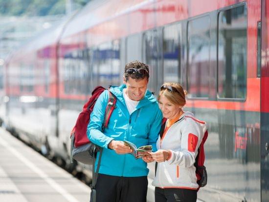 Smiling couple with backpacks reading a brochure at the train station beside a red regional train