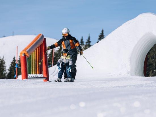 Adult teaching child to ski on snowy slope with colorful obstacle and snow tunnel