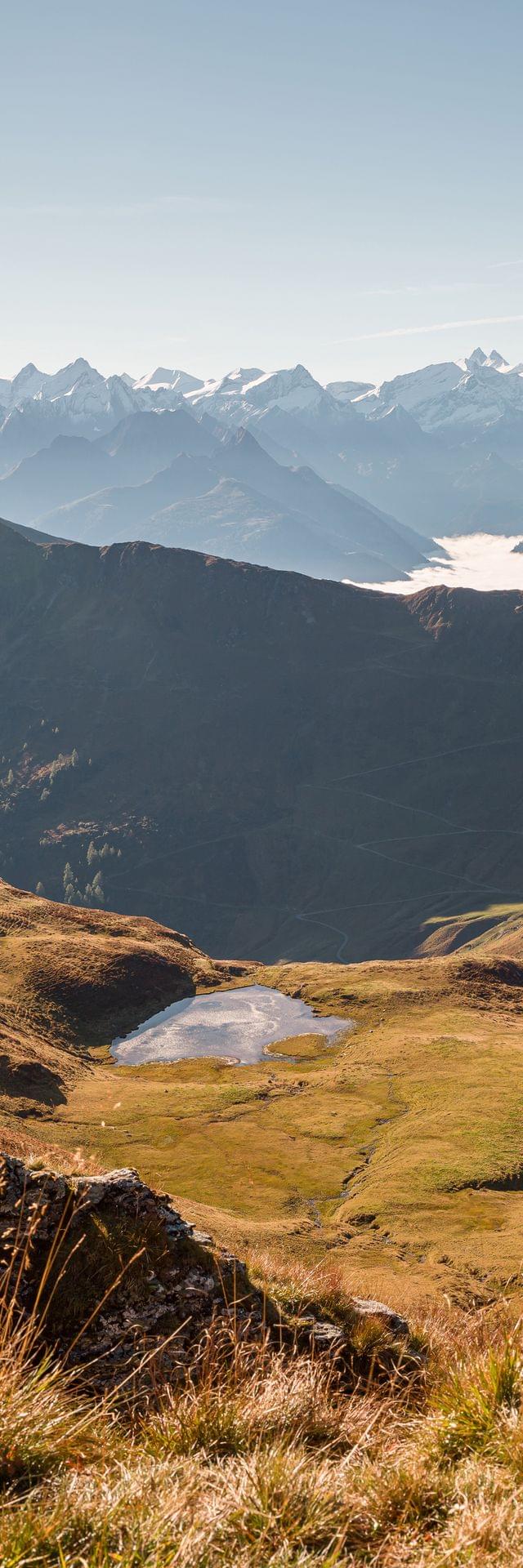 Alpine Landschaft mit grasbewachsenen Hängen, kleinem See und schneebedeckten Bergen im Hintergrund