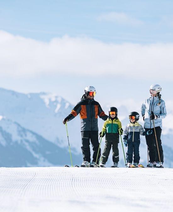 Family skiing on a groomed slope with snowy mountain peaks in the background