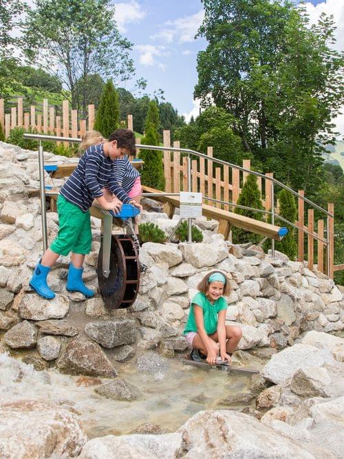 Children playing at a water play area with rocks and a small water wheel in a mountain park