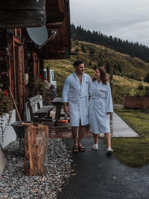 Two people in bathrobes in front of an alpine hut, with an autumnal atmosphere in the background.