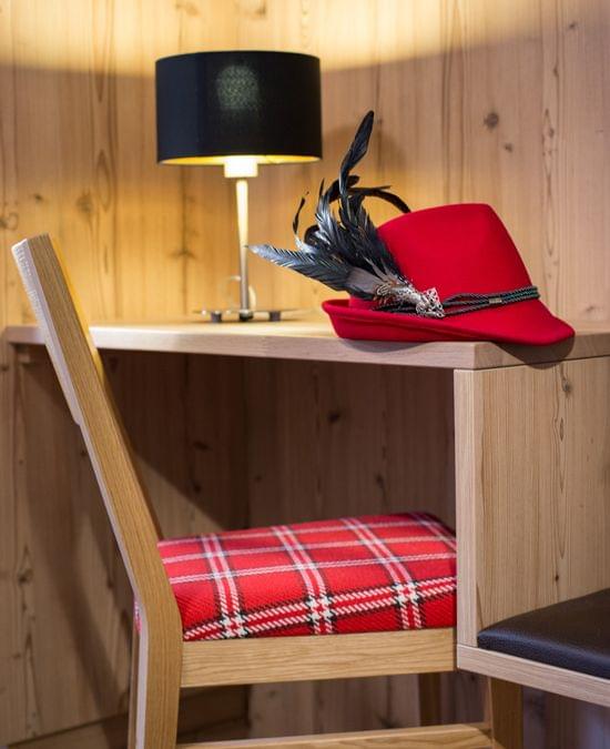 Roter traditioneller Alpenhut mit Federn auf Holzschreibtisch neben kariertem Stuhl